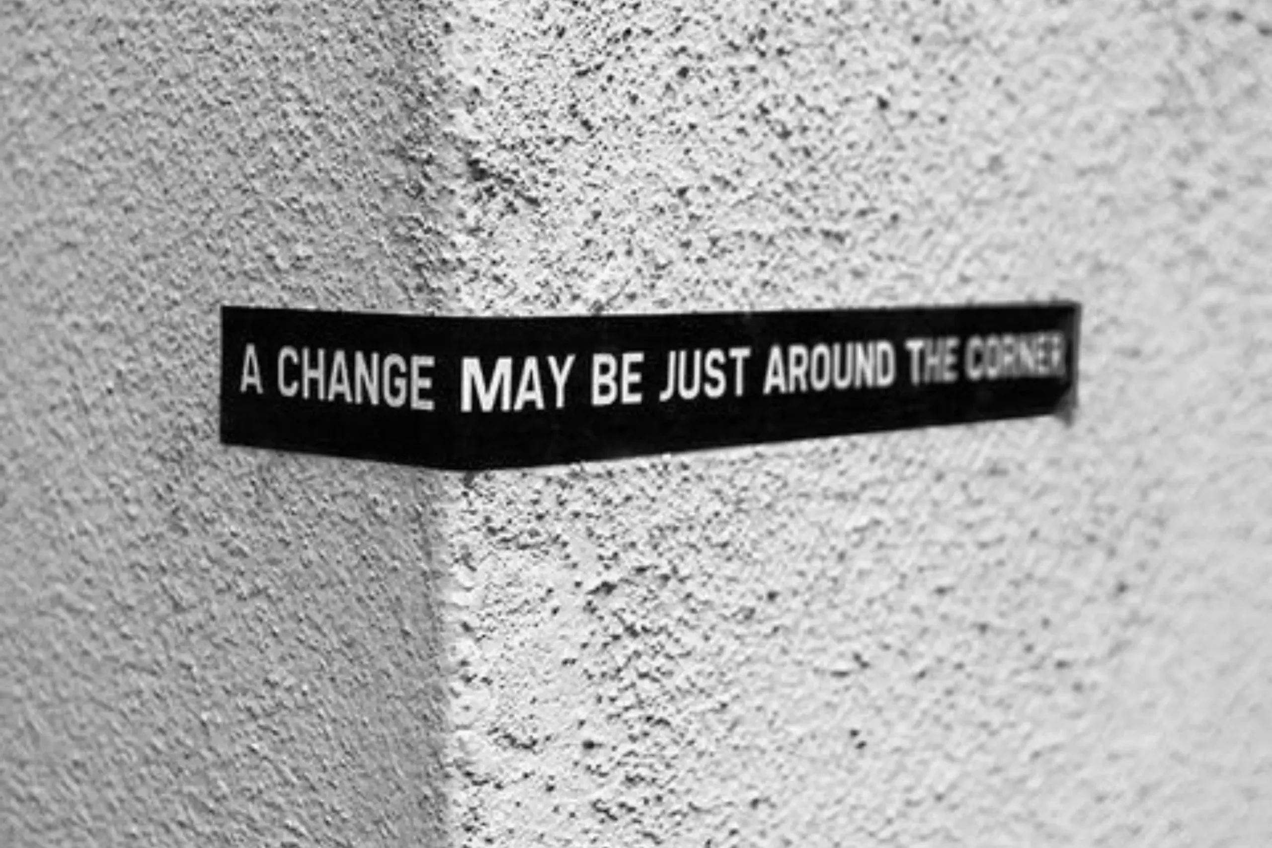 A black sign on a white wall reading 'A change may be just around the corner"